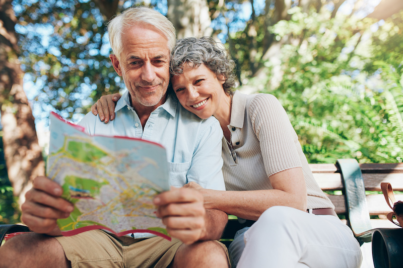 Couple of mature tourist on a vacation. Woman embracing man holding a map. Happy senior couple relaxing on a park bench reading a city map.